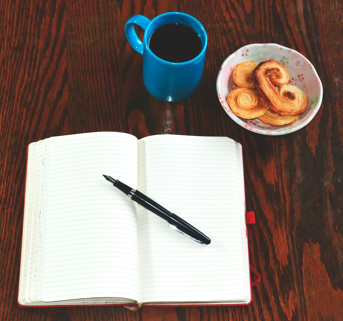 Offener Notizblock mit Stift, Kaffeetasse und Gebäck auf einem Holztisch.